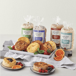 A selection of Wolferman's Bakery super-thick English muffins in flavors like San Francisco Sourdough, Wild Maine Blueberry, Cinnamon Raisin, and 1910 Original Recipe, displayed with fresh berries and orange slices.
