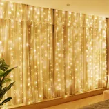 Warm white LED string lights draped over sheer curtains create a cozy, illuminated backdrop. A plant is partially visible on the left.