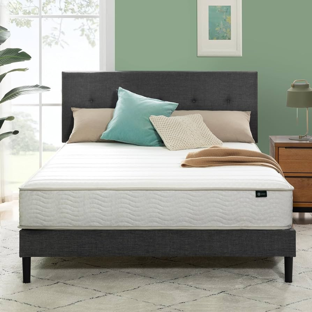 A neatly made bed with a minimalistic white mattress featuring a wave pattern, placed on a dark upholstered bed frame. The setting includes neutral and teal pillows and a wooden nightstand with a lamp.
