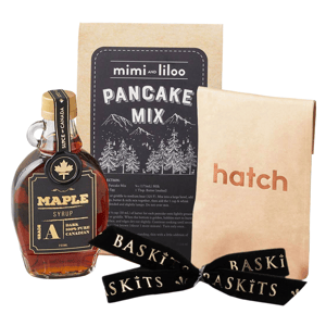 Pancake mix from \“mimi and liloo,\“ maple syrup labeled \“Grade A Dark 100% Pure Canadian\“ with a leaf emblem, and a package labeled \“hatch,\“ all tied with a \“Baskits\“ ribbon.