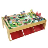 A wooden train set table featuring a complete cityscape with tracks, bridges, buildings, vehicles, and figures, including three red storage bins underneath.