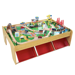 A wooden train set table featuring a complete cityscape with tracks, bridges, buildings, vehicles, and figures, including three red storage bins underneath.