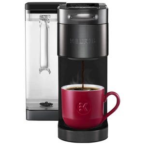 A Keurig single-serve coffee maker with a modern design is shown brewing coffee into a red mug. It features a water reservoir on the side and a sleek, compact form.