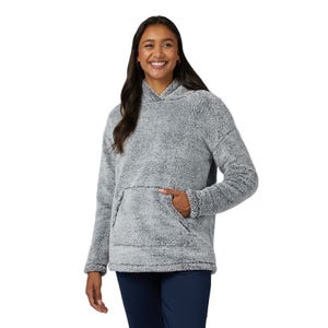 A woman is wearing a light gray, long-sleeve Sherpa pullover with a hood and a front kangaroo pocket, paired with navy blue pants. The pullover has a soft, plush texture ideal for warmth and comfort.