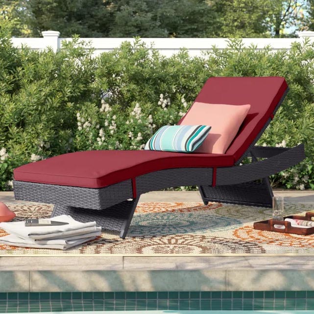 A rattan outdoor chaise lounge with red cushions is positioned by a pool, accompanied by decorative throw pillows in striped and solid patterns, and a colorful outdoor rug.
