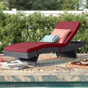 A rattan outdoor chaise lounge with red cushions is positioned by a pool, accompanied by decorative throw pillows in striped and solid patterns, and a colorful outdoor rug.