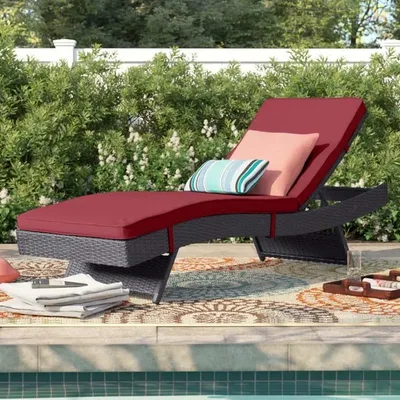 A rattan outdoor chaise lounge with red cushions is positioned by a pool, accompanied by decorative throw pillows in striped and solid patterns, and a colorful outdoor rug.