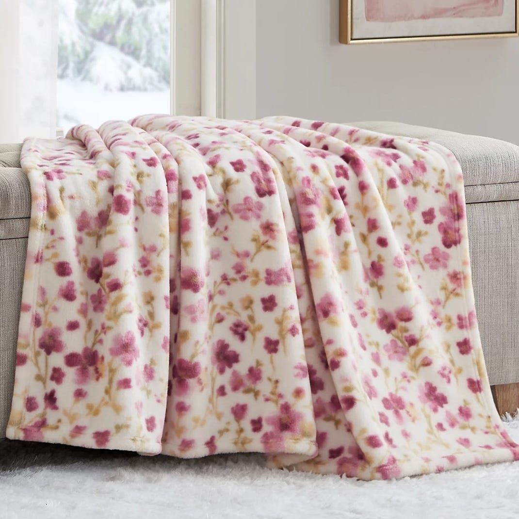 A soft, oversized plush throw with a cream background, featuring a floral pattern in shades of pink and brown. It's draped elegantly over a beige ottoman, suggesting comfort and warmth.