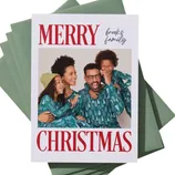 A Christmas card features a family photo with \“Merry Christmas\“ in red text and the words \“Brooks family\“ handwritten above. The card is placed on a stack of green envelopes against a green background.