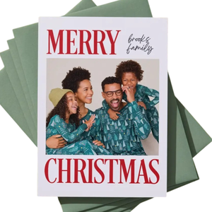 A Christmas card features a family photo with \“Merry Christmas\“ in red text and the words \“Brooks family\“ handwritten above. The card is placed on a stack of green envelopes against a green background.