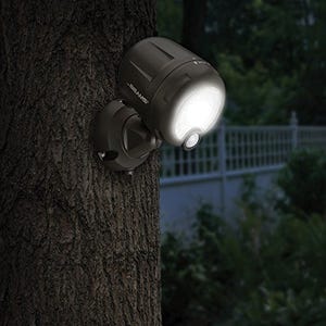 A battery-powered LED spotlight is mounted on a tree, featuring a motion sensor and adjustable head for targeted illumination. The light is on, suggesting dusk or nighttime conditions.