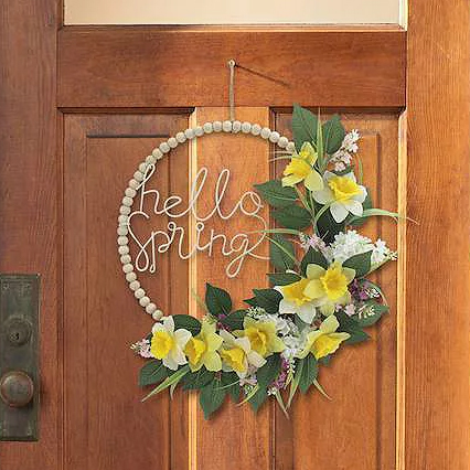 A wooden door features a circular wreath with the words \“hello spring\“ and decorated with yellow and white flowers and green leaves.