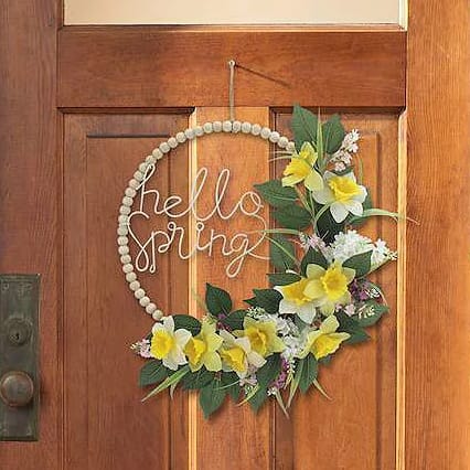 A wooden door features a circular wreath with the words \“hello spring\“ and decorated with yellow and white flowers and green leaves.
