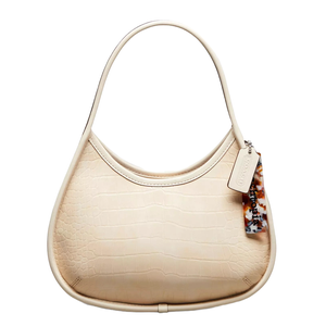 Cream-colored bag with a crocodile-embossed texture, featuring a curved, hobo-style shape. It has a single strap, a tortoiseshell charm, and a Coach tag attached.