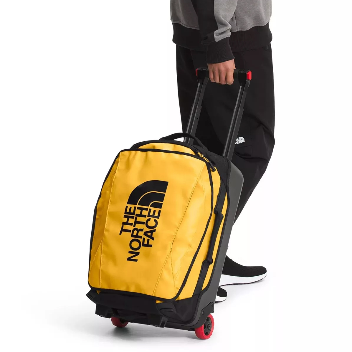 A person is rolling a yellow wheeled suitcase with a prominent logo. The suitcase has a telescoping handle and red wheel accents.