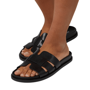 A pair of black, slip-on sandals with wide straps and a flat sole, worn by a person with a light brown skin tone.