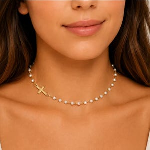 A woman wears a pearl choker necklace featuring a small horizontal gold cross.