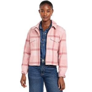 A woman is wearing a pink and white plaid relaxed shirt jacket with front pockets over a denim outfit.