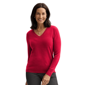 A woman wears a red V-neck sweater with long sleeves and black patterned pants, accessorized with a gold necklace and earrings, against a plain background.