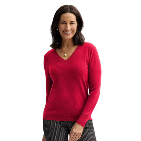 A woman wears a red V-neck sweater with long sleeves and black patterned pants, accessorized with a gold necklace and earrings, against a plain background.