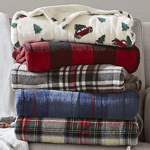 A neatly folded stack of five blankets sits on a couch, featuring various designs: white with trees and red cars, red and black checkered, brown plaid, solid blue, and multicolored tartan plaid.