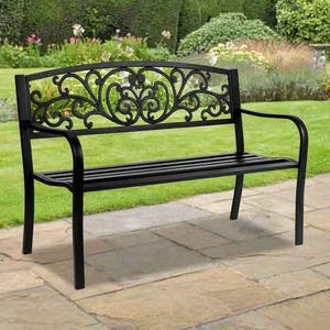 A black metal garden bench with ornate scrollwork on the backrest is placed on a stone patio, set against a landscaped garden background.