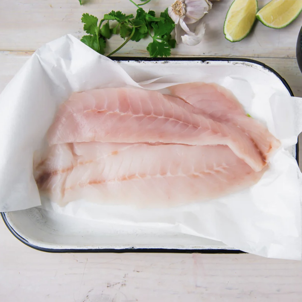 Raw fish fillets on a tray with cilantro, lime wedges, garlic, and a bowl of pickled red onions.