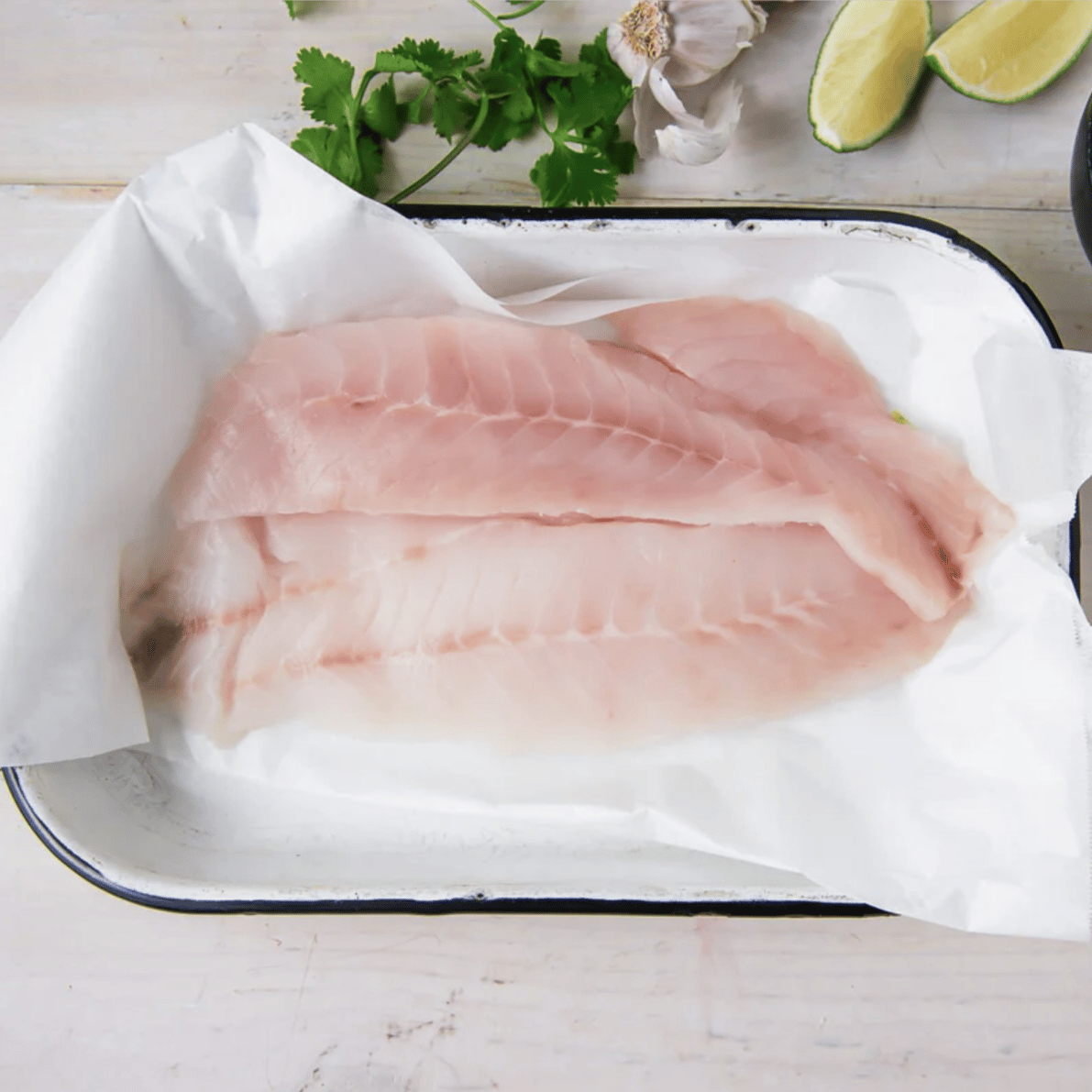 Raw fish fillets on a tray with cilantro, lime wedges, garlic, and a bowl of pickled red onions.