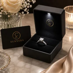 A diamond engagement ring is showcased in an open black box with an LED light, accompanied by a card bearing the name \“Sunnera.\“ The setting includes a vase with a white rose and small flowers, with a candle in the background.