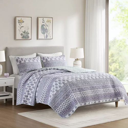 A bedroom features a neatly made bed with a patterned duvet and matching pillows, flanked by two white nightstands with lamps. Two floral prints are hung above the headboard, and a window allows natural light to fill the room.