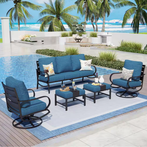 Outdoor 7-seat patio set with blue cushions includes a sofa, two armchairs, two ottomans, and two coffee tables on a blue-bordered rug, set in a poolside environment with palm trees and ocean in the background.