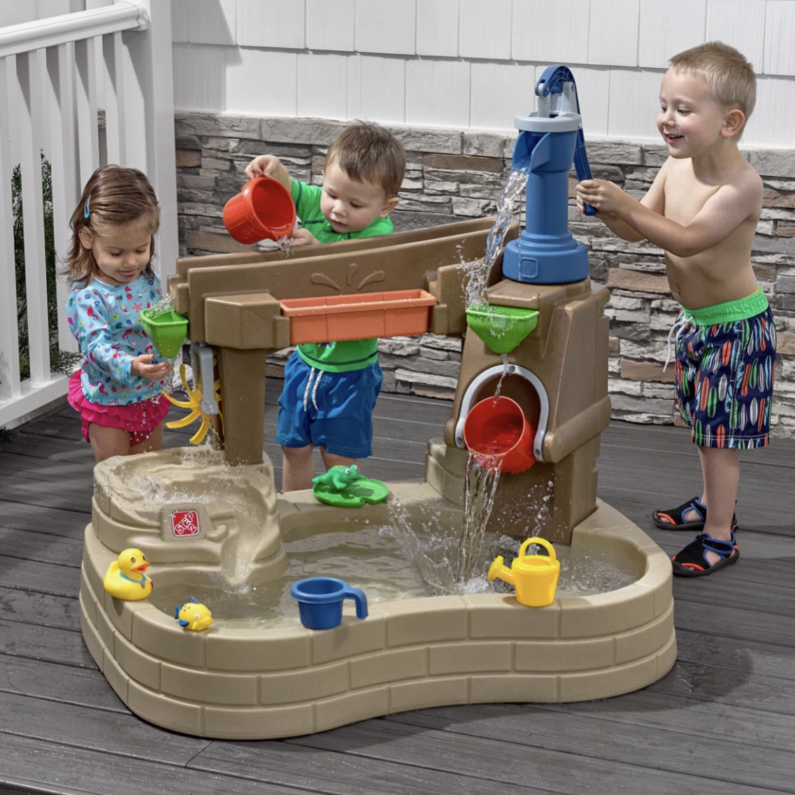 Children playing with a water activity table featuring a pump, buckets, and various water toys like rubber ducks and watering cans, designed for outdoor fun.