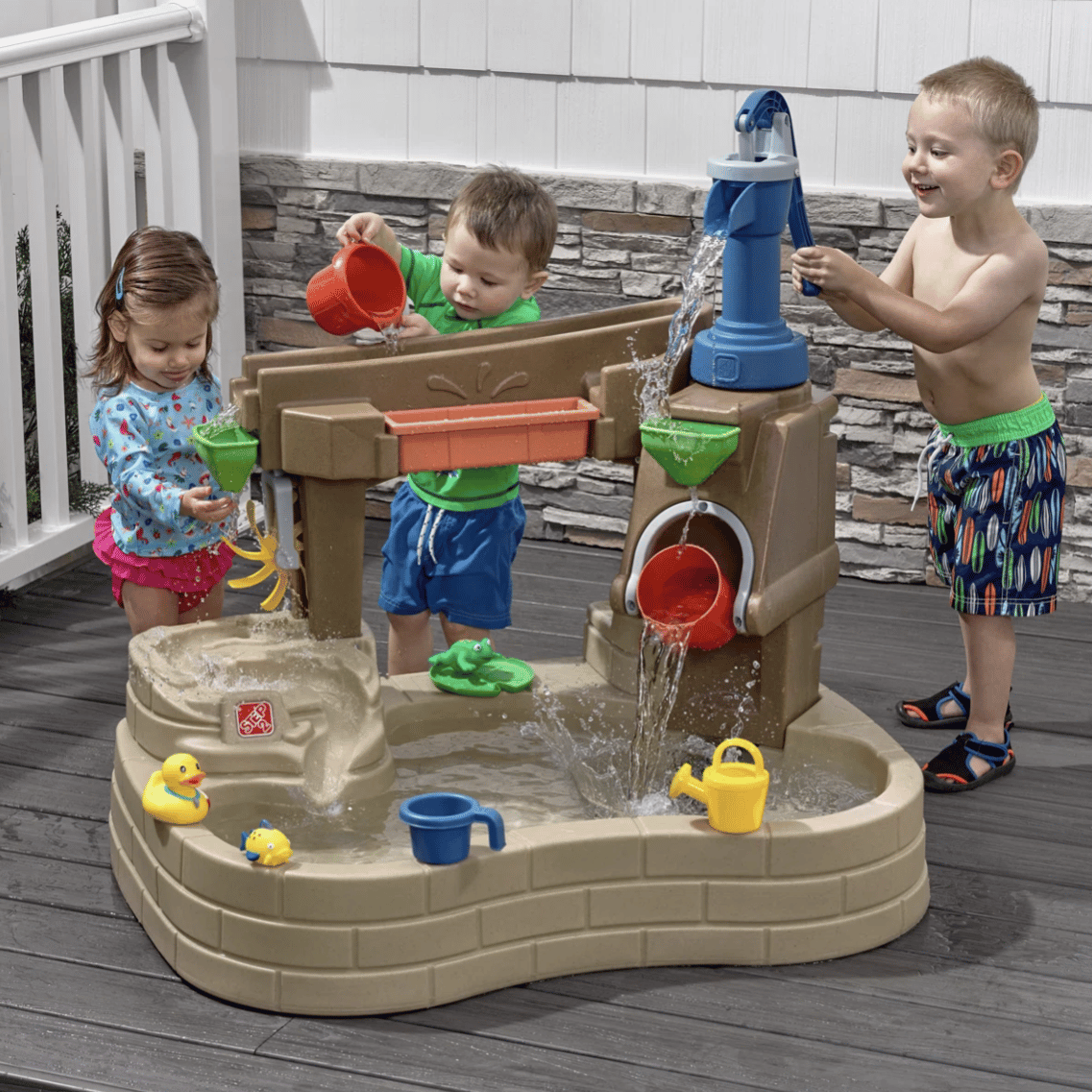 Children playing with a water activity table featuring a pump, buckets, and various water toys like rubber ducks and watering cans, designed for outdoor fun.