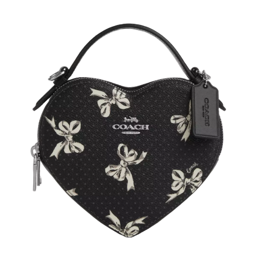 The Coach Heart Crossbody Bag features a black heart-shaped design with a white bow print, zip closure, and top handle. It includes a Coach tag and logo on the front.
