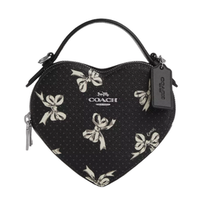 The Coach Heart Crossbody Bag features a black heart-shaped design with a white bow print, zip closure, and top handle. It includes a Coach tag and logo on the front.