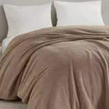 A beige, textured blanket is neatly spread over a bed with two white pillows against a light gray wall.