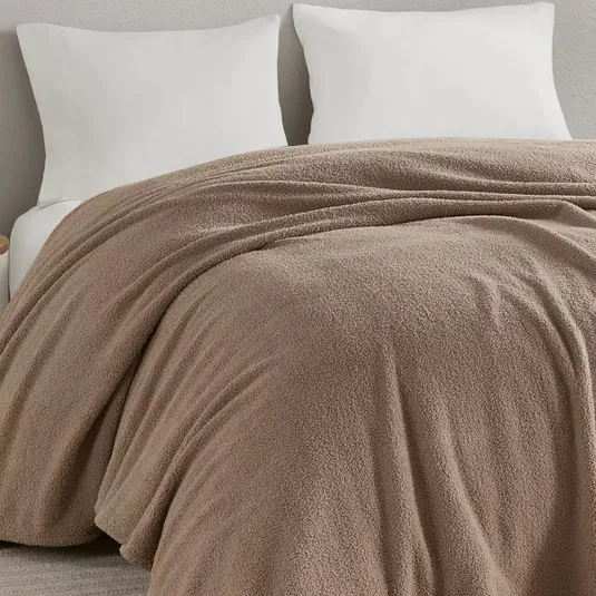 A beige, textured blanket is neatly spread over a bed with two white pillows against a light gray wall.