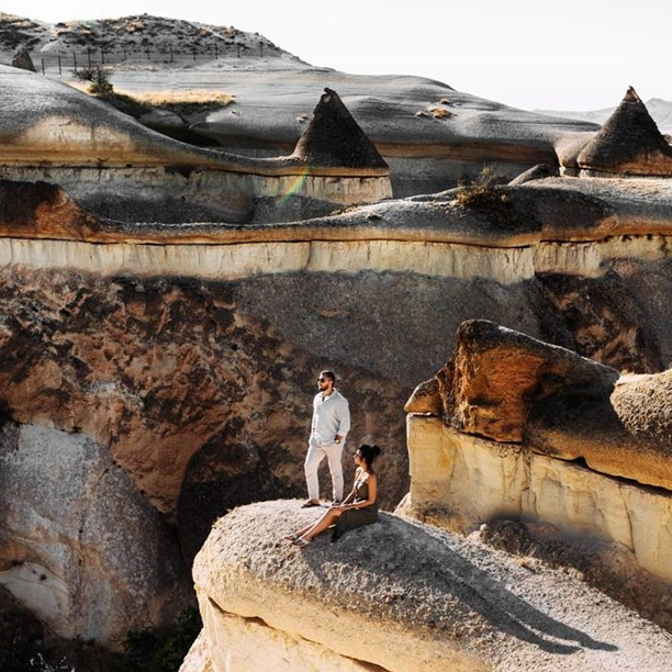A man and a woman are on rock formations, possibly in a scenic, arid location with unique, conical structures.