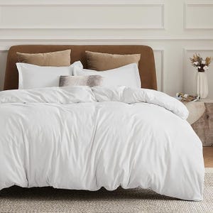 A white comforter set with matching pillows on a bed featuring a brown headboard, accented with beige and light gray pillows in a bedroom setting.