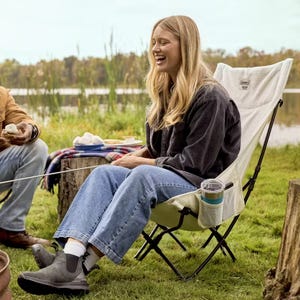 A person is seated on a Coleman camping chair with a cup holder and pocket, laughing during an outdoor marshmallow roasting session by a lake.