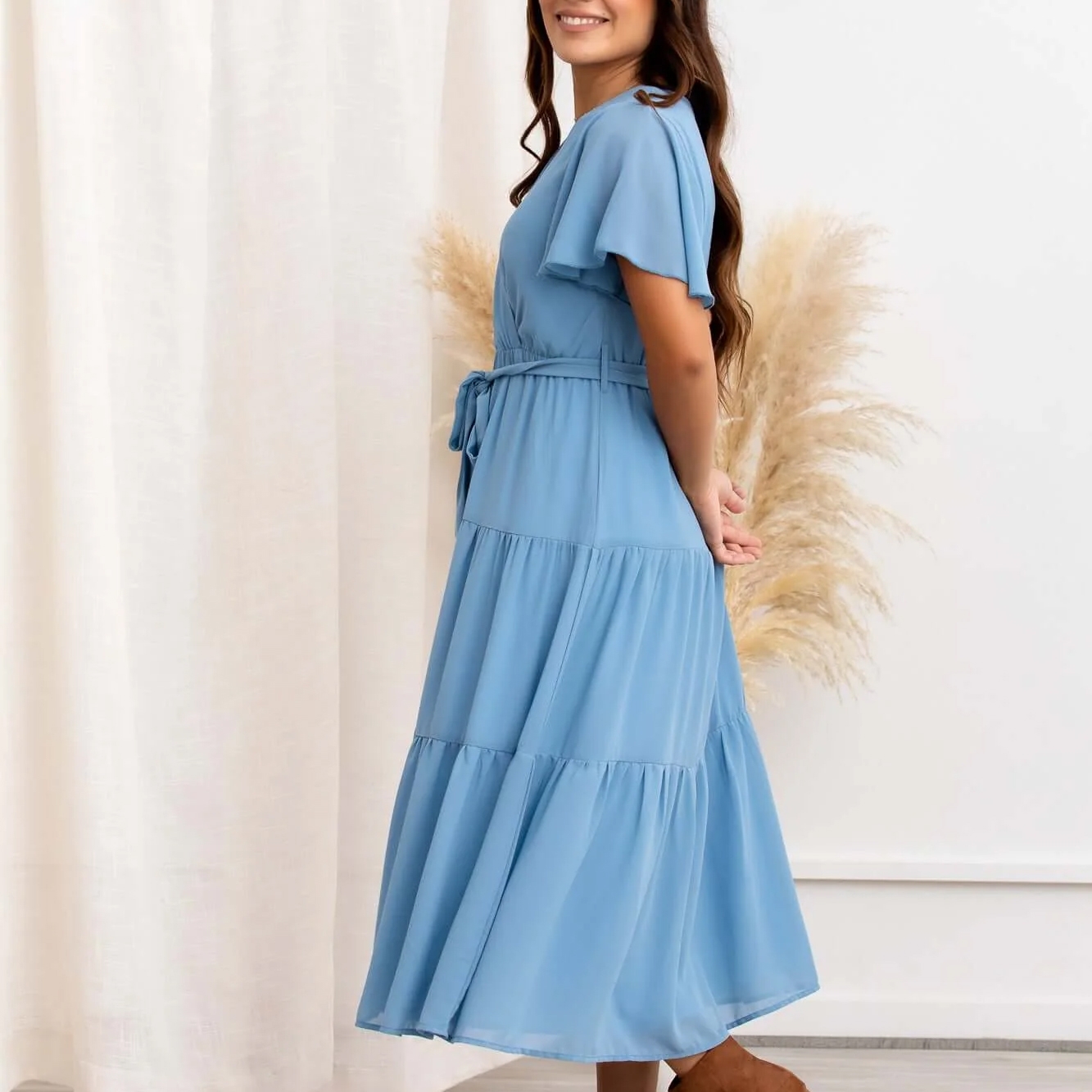 A woman is wearing a light blue, tiered midi dress with short ruffled sleeves and a waist tie, standing against a neutral backdrop.