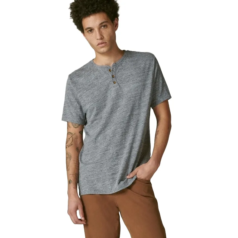 A man is wearing a heather gray short-sleeve linen shirt with a partial button-up neckline and visible tattoos on his left arm. He pairs it with brown trousers.