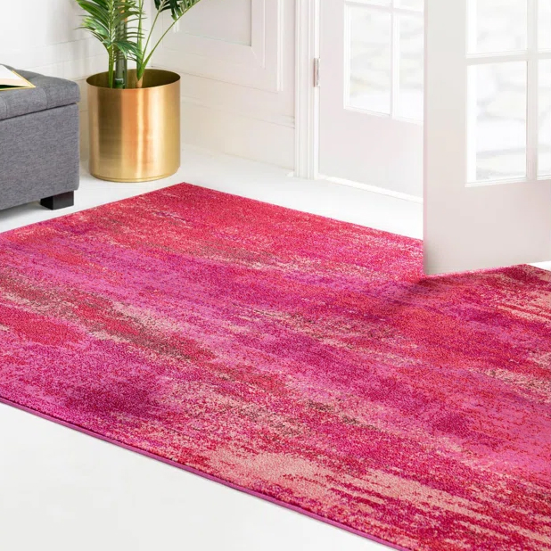 A vibrant red and pink area rug is displayed on a white floor, positioned near a grey sofa and a brass planter with a plant, adjacent to a glass-paneled door.