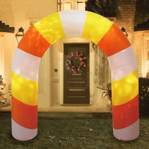 An inflatable arch with yellow, orange, and white stripes is set up in front of a house, resembling a piece of candy corn. A decorative wreath is visible on the door behind it.