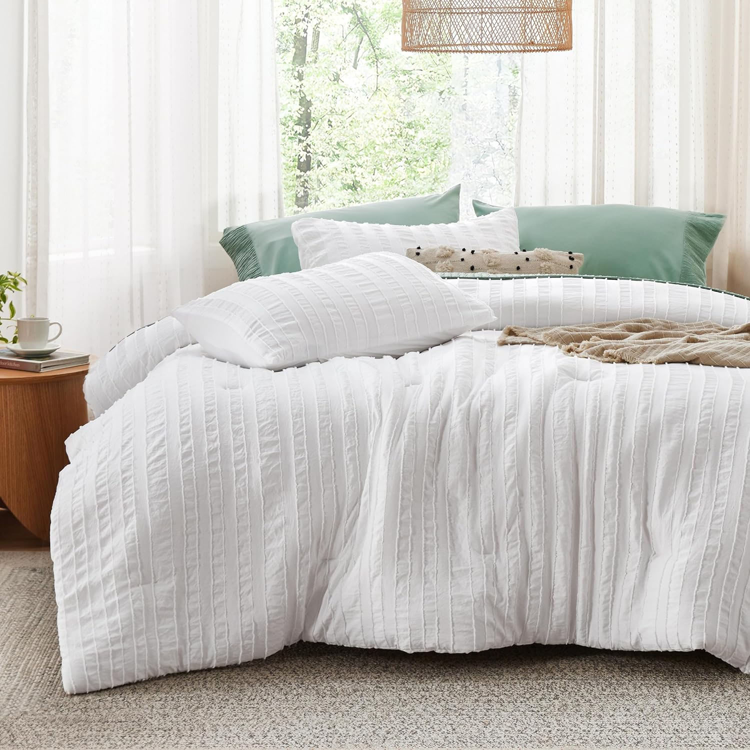White, textured Queen Farmhouse Comforter Set featuring a subtle striped design, paired with matching pillow shams, and styled with green accent pillows and a beige throw in a bright, airy bedroom setting.