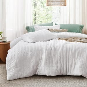 White, textured Queen Farmhouse Comforter Set featuring a subtle striped design, paired with matching pillow shams, and styled with green accent pillows and a beige throw in a bright, airy bedroom setting.