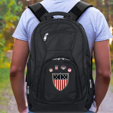 A person wearing a grey t-shirt is carrying a black backpack with an American flag badge and various pins.