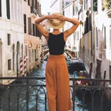 A person holds a straw hat while standing on a bridge overlooking a canal. They wear a black sleeveless top and rust-colored wide-leg pants.