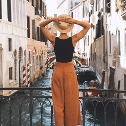 A person holds a straw hat while standing on a bridge overlooking a canal. They wear a black sleeveless top and rust-colored wide-leg pants.