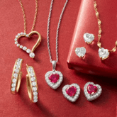 Assorted jewelry on a red background, featuring heart-shaped pendants with gemstones, gold hoop earrings with diamonds, and matching heart earrings with a red gift box.